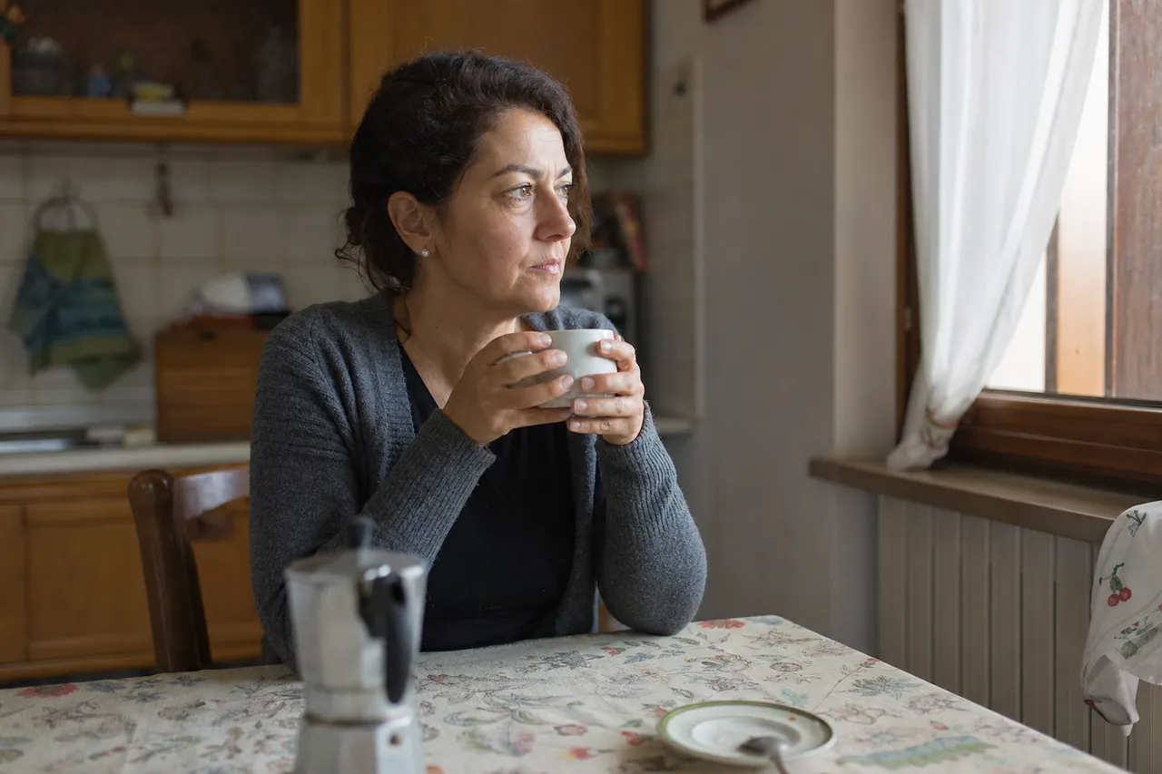 Una donna adulta si concede una pausa seduta al tavolo di casa, con una tazza tra le mani e lo sguardo assorto